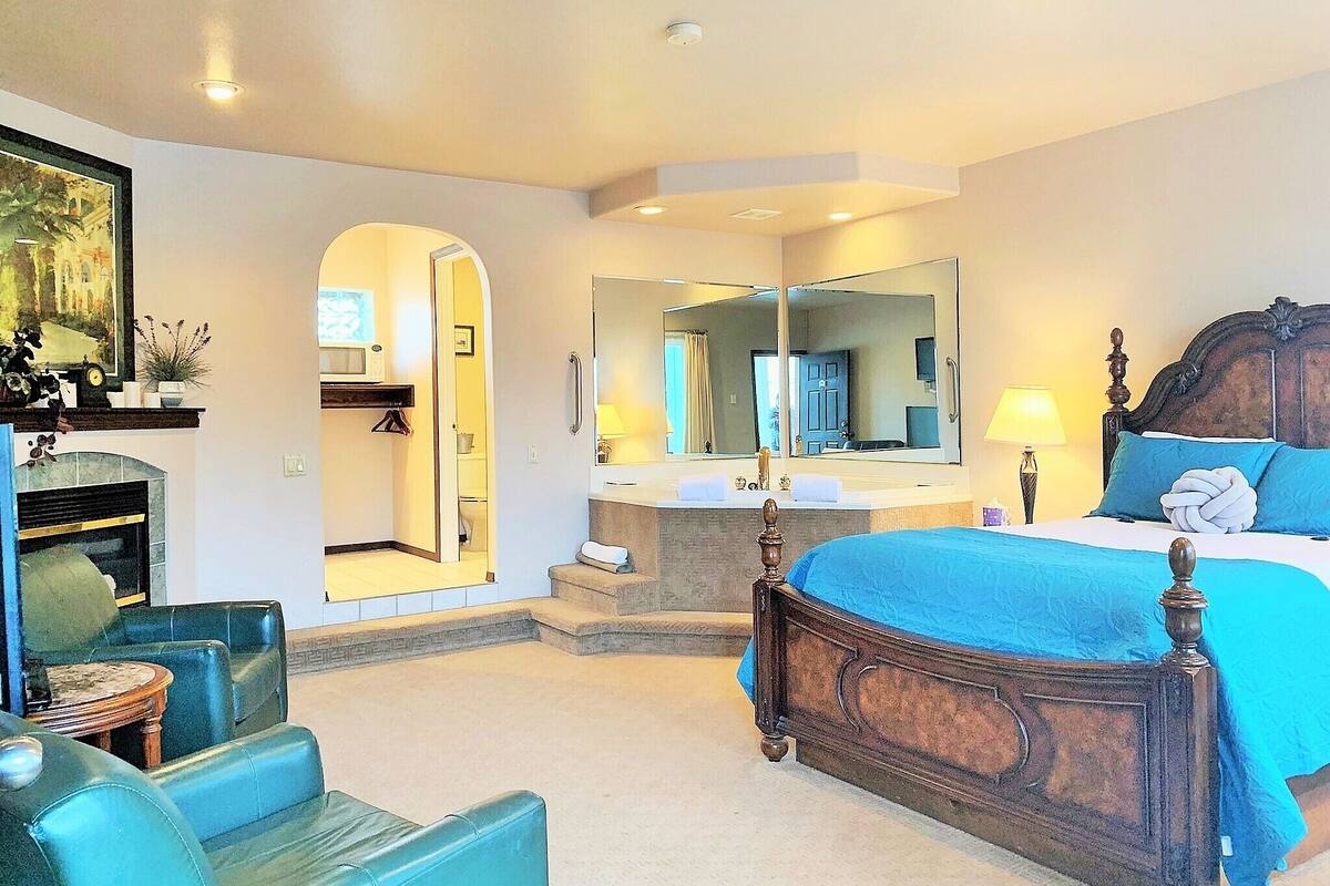 A bedroom with a large bed, a sitting area with chairs, a fireplace, a mirror, and a bathroom visible through an archway.