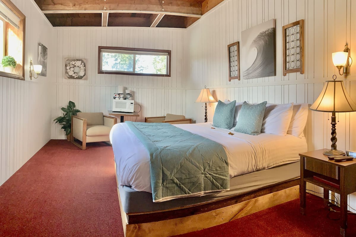 A bedroom with a wooden ceiling, a bed with a green quilt, two bedside lamps, a small table with a microwave, and a window with a view of trees.
