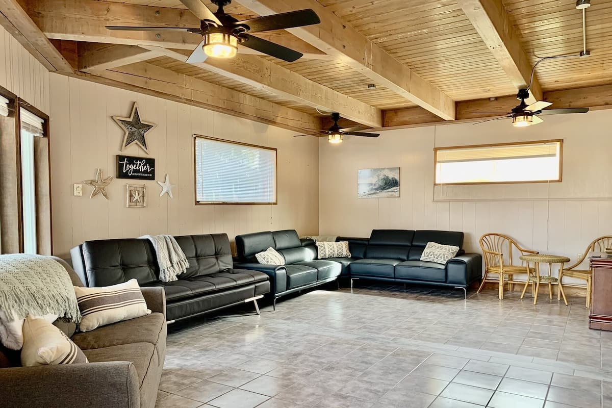 A spacious living area with a high ceiling, wooden beams, and ceiling fans. There are two black leather sofas with patterned cushions, a grey armchair, and a small round table with wicker chairs. The floor is tiled, and there are star-shaped wall decorations and a framed picture with text.