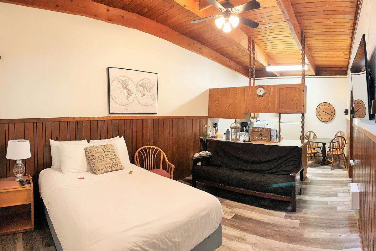 A hotel room with a bed, a wooden headboard, a small table with a lamp, a chair, a sofa, a clock, and a wall-mounted TV.