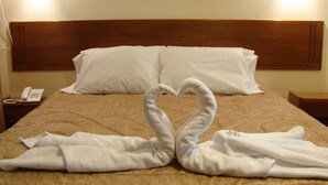 Quarto de hotel com uma cama, cabeceira de madeira, almofadas brancas, telefone ao lado da cama e duas toalhas dobradas em forma de coração.