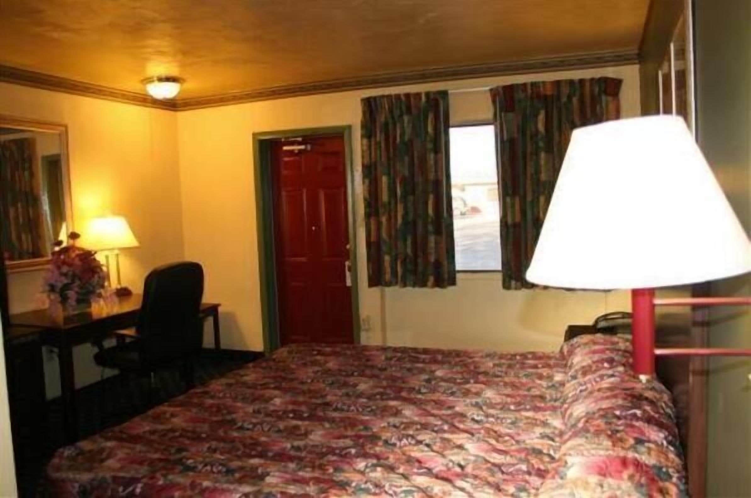 Ein Hotelzimmer mit einem Bett, einem Schreibtisch mit Lampe, einem Sessel und einer roten Tür.