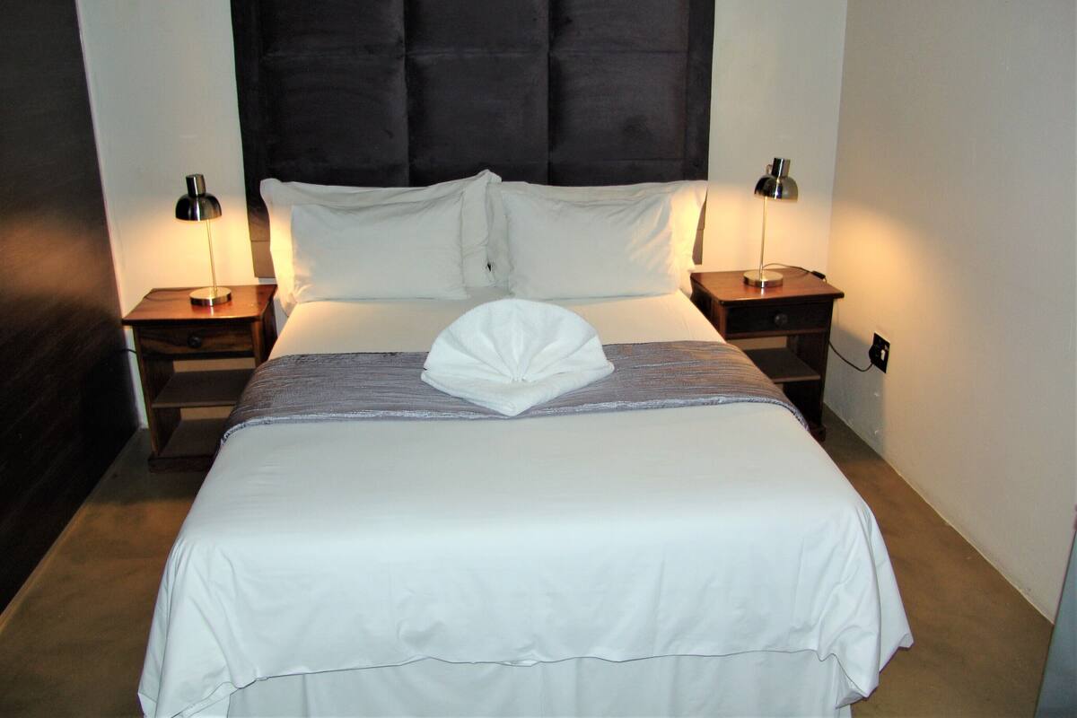 A neatly made bed with a folded towel, two bedside tables with lamps, and a dark headboard.