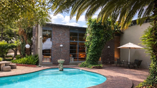 A swimming pool with a fountain, surrounded by a brick patio, with a building featuring large windows and a brick facade in the background.