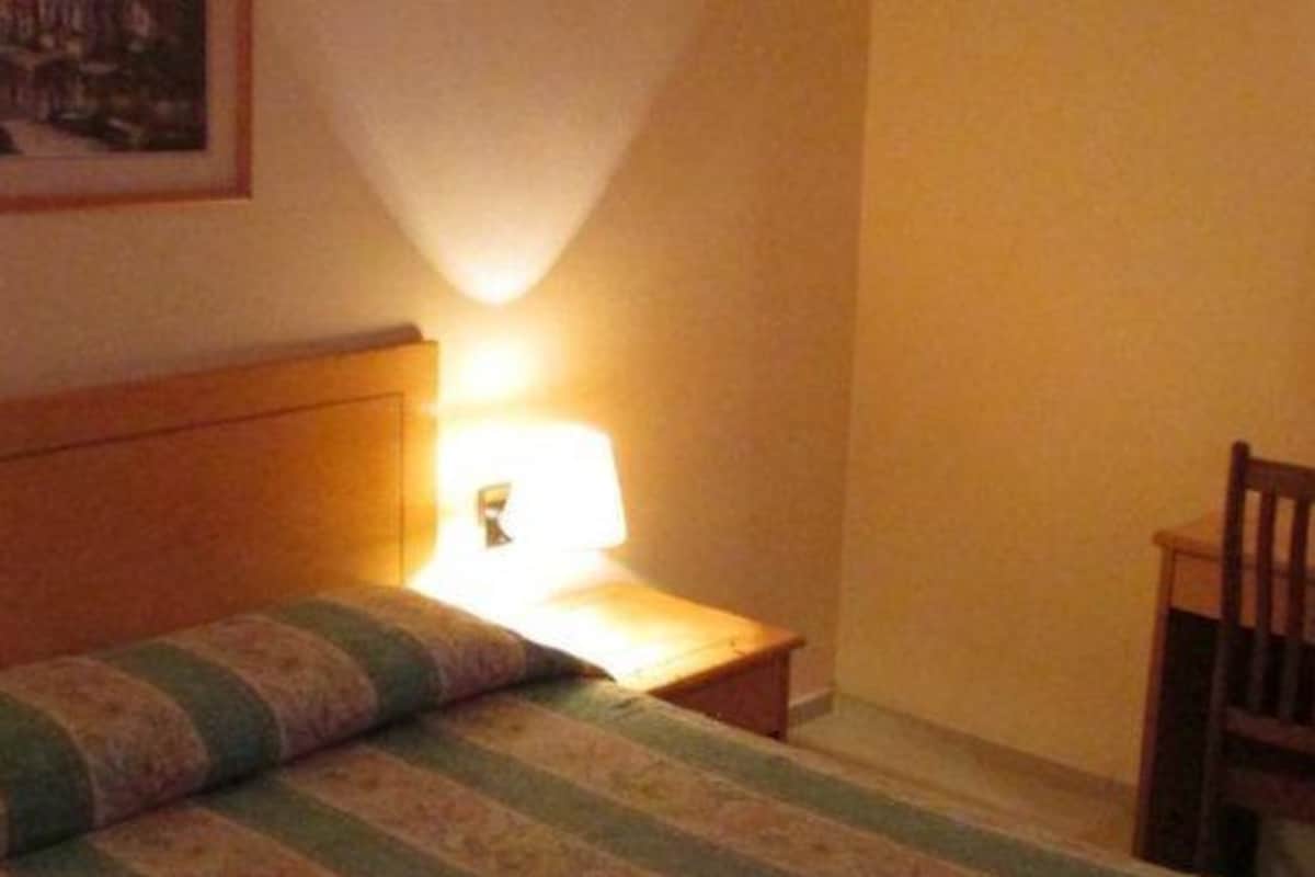 A small, cozy room with a single bed, a nightstand with a lamp, and a chair.