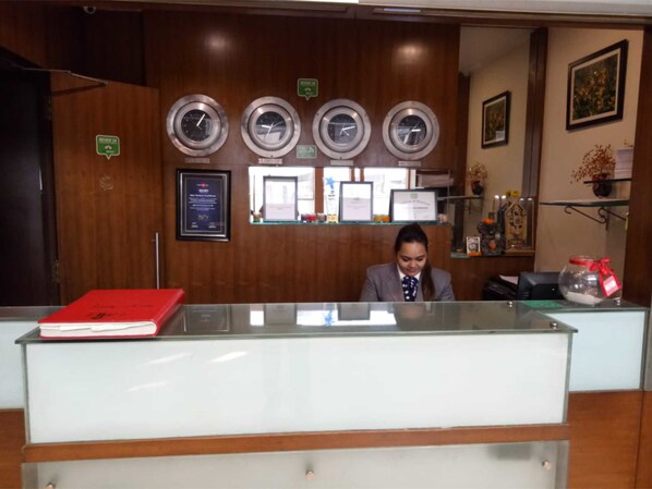 Un mostrador de recepción con una superficie de cristal, un libro rojo encima y una mujer sentada detrás. Hay cuatro relojes en la pared sobre el mostrador.