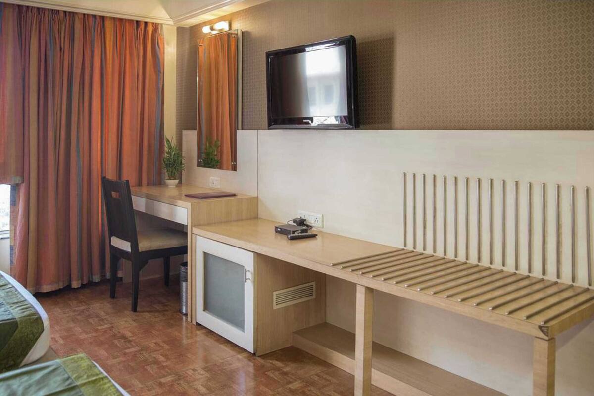 A compact hotel room with a wooden desk, a small refrigerator, a television mounted above the desk, and a window with curtains.