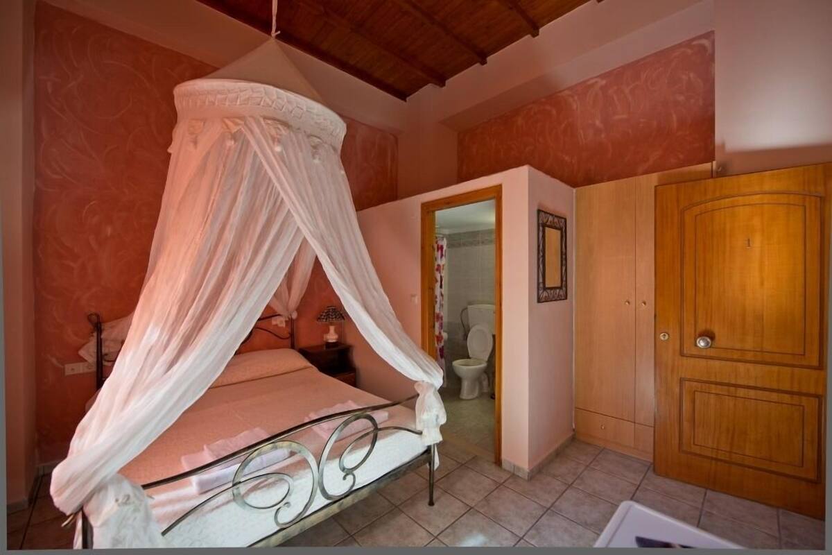 Een slaapkamer met een hemelbed, een houten deur en een zichtbare badkamer door een open deuropening.