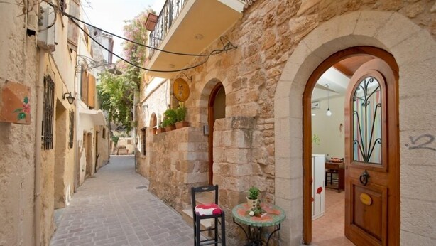 A narrow alleyway with stone buildings, a wooden door with a decorative glass panel, and a small round table with a chair and a potted plant.