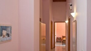 A narrow hallway with pink walls, wooden ceiling beams, and a tiled floor. There is a small table with a lamp and a framed picture on the left.