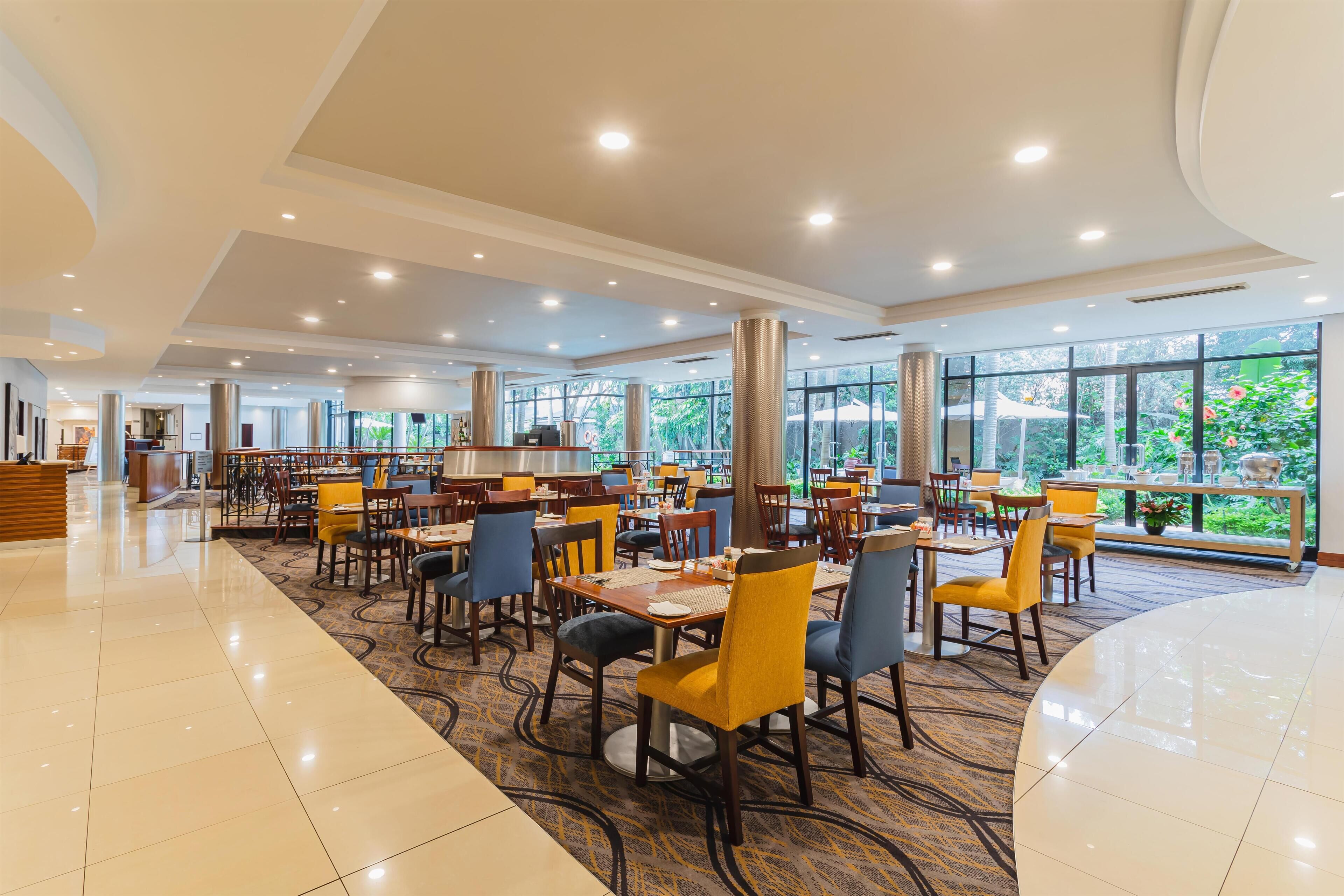 A spacious dining area with multiple tables and chairs, large windows offering a view of greenery, and a clean, modern interior design.