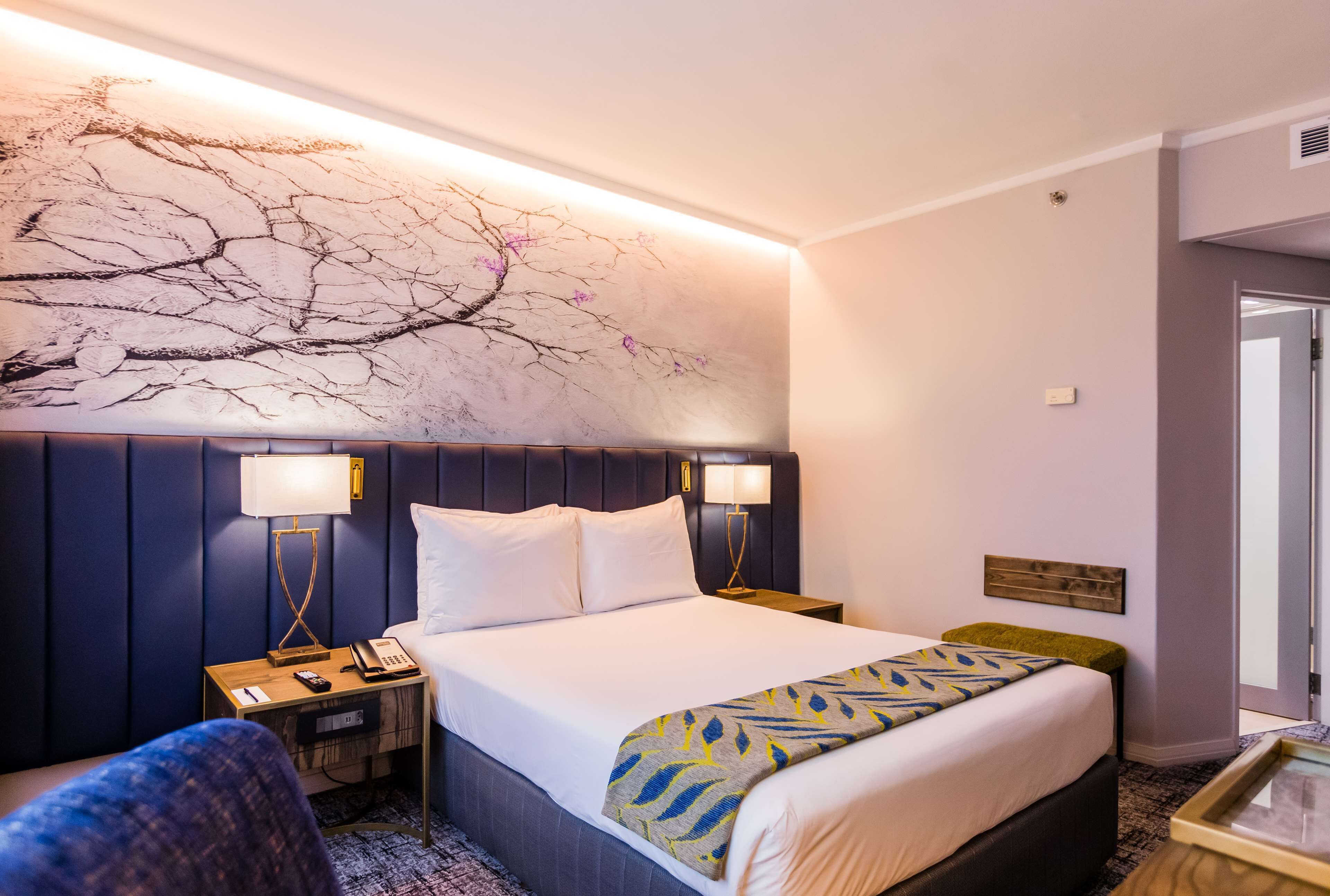A hotel room with a large bed, two bedside tables with lamps, a nightstand, a chair, and a wall-mounted artwork.