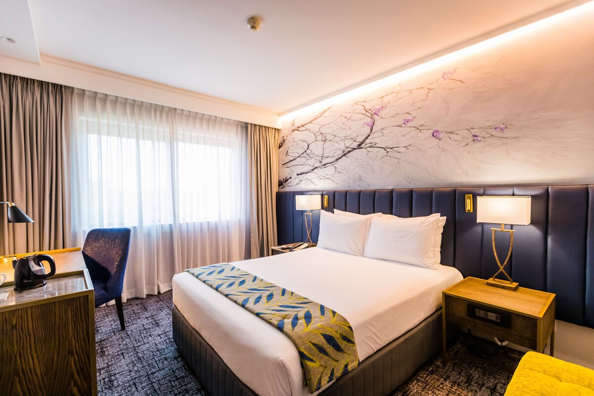 A hotel room with a large bed, a desk, a chair, and a nightstand with lamps.
