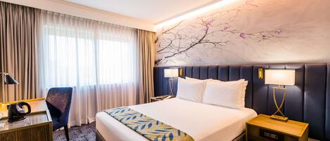A hotel room with a large bed, a desk, a chair, and a nightstand with lamps.