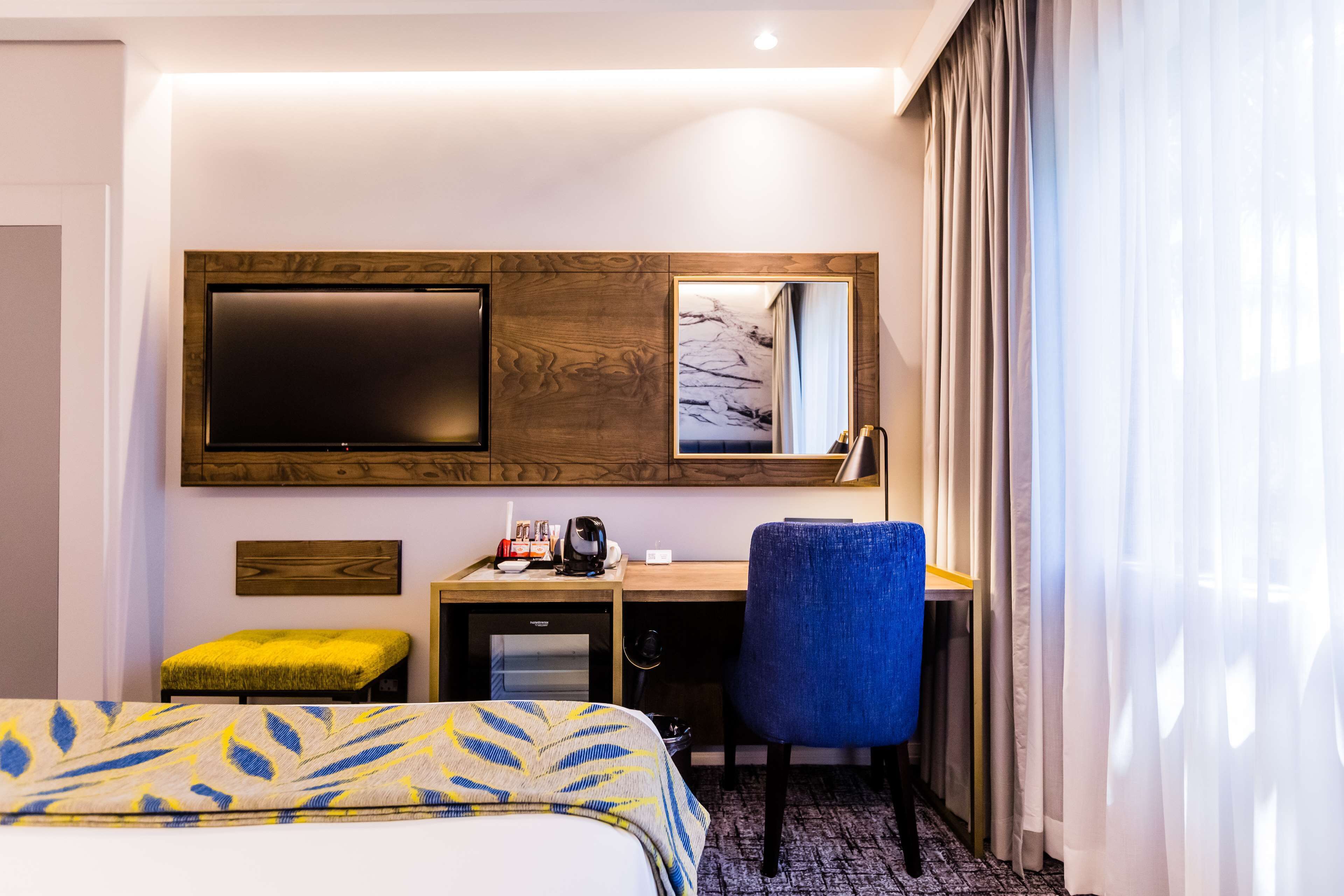 A hotel room with a bed, a desk with a chair, a television, a mirror, and a window with curtains.