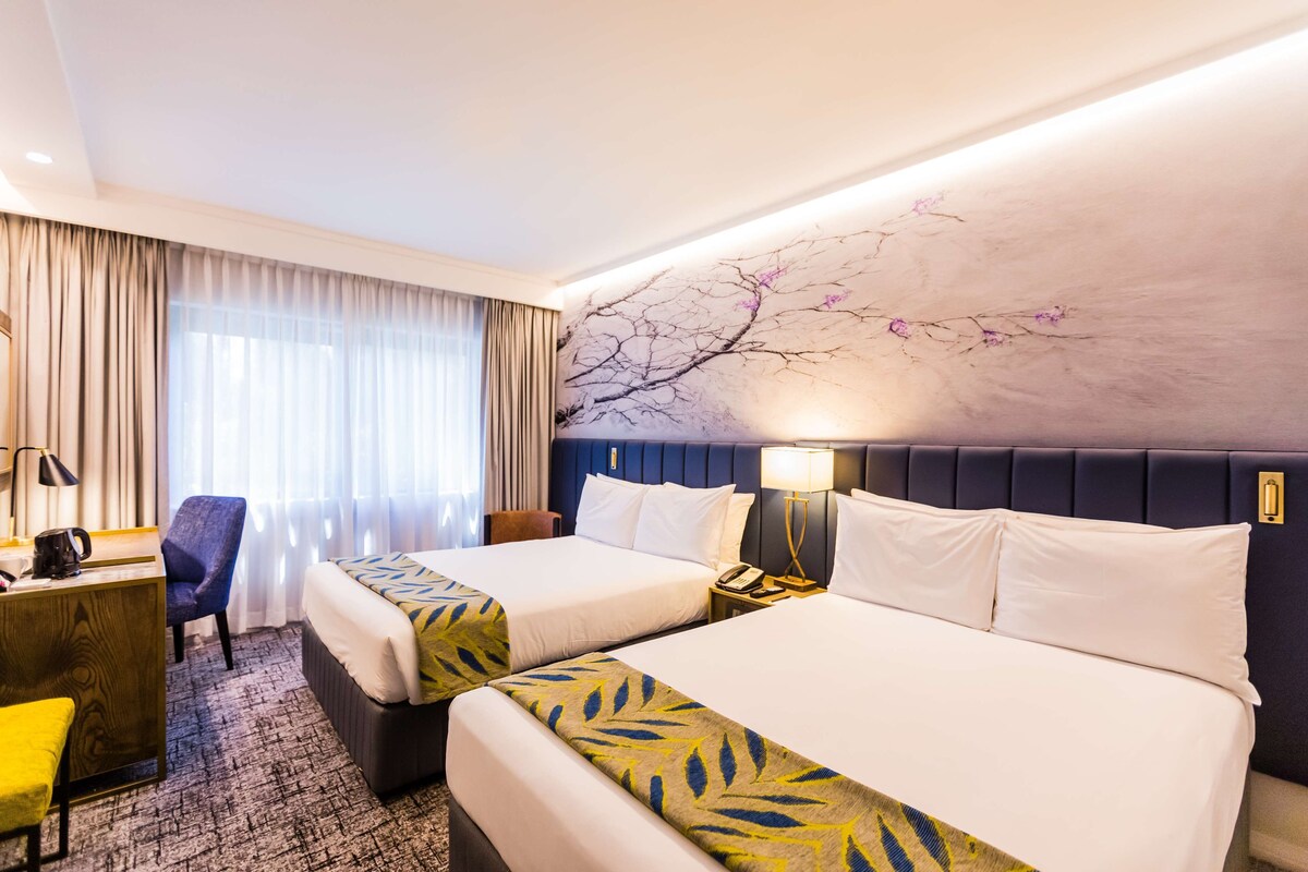 A hotel room with two beds, a desk, a chair, and a large window with curtains.