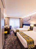Habitación de hotel con dos camas, un escritorio, una silla y un ventanal con cortinas.