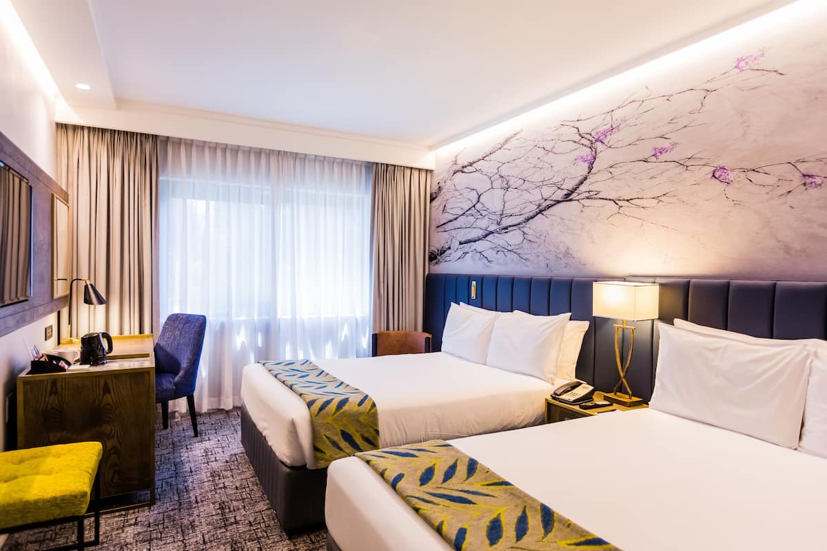A hotel room with two beds, a desk, a chair, a desk lamp, and a wall with a tree mural.