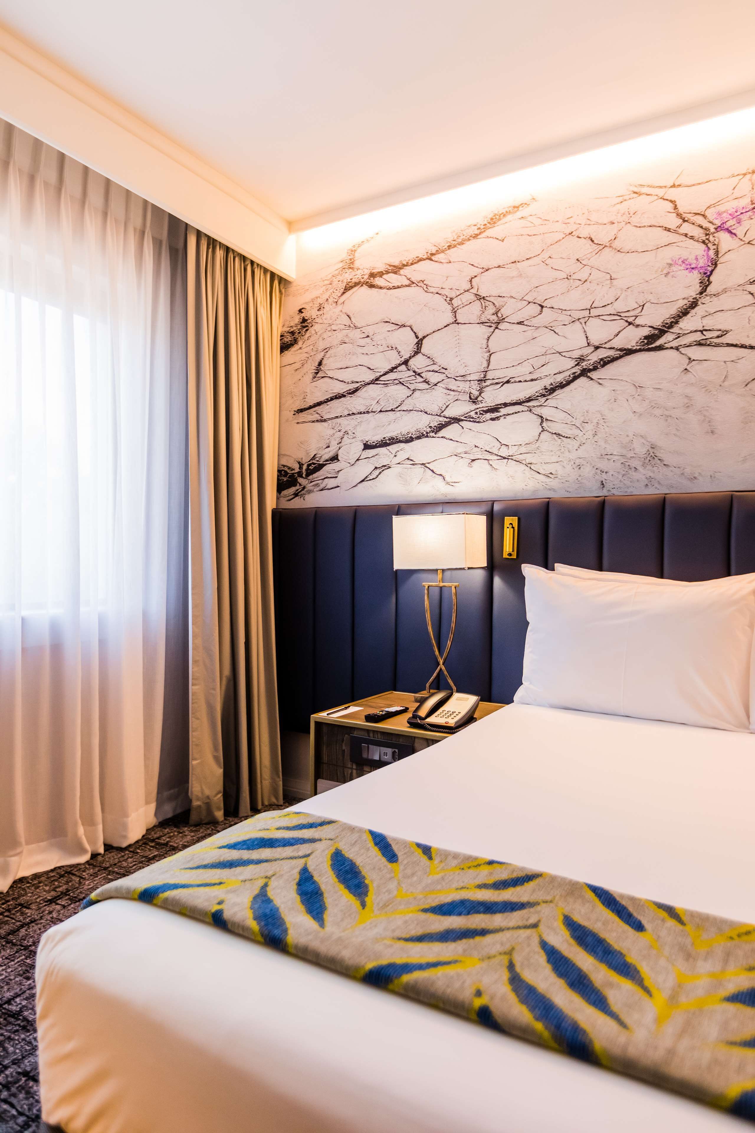 Habitación de hotel con una cama grande, una mesita de noche con lámpara y una pared con un mural de una rama de árbol.
