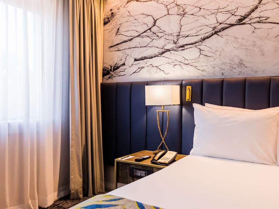 Habitación de hotel con una cama grande, una mesita de noche con lámpara y una pared con un mural de una rama de árbol.