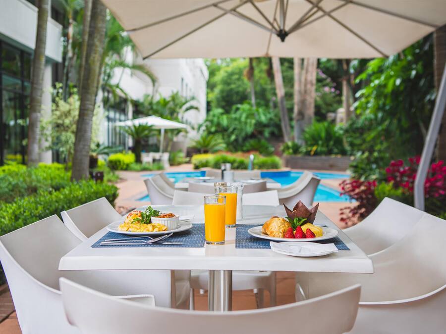 A dining area with white tables and chairs, a pool in the background, and lush greenery.