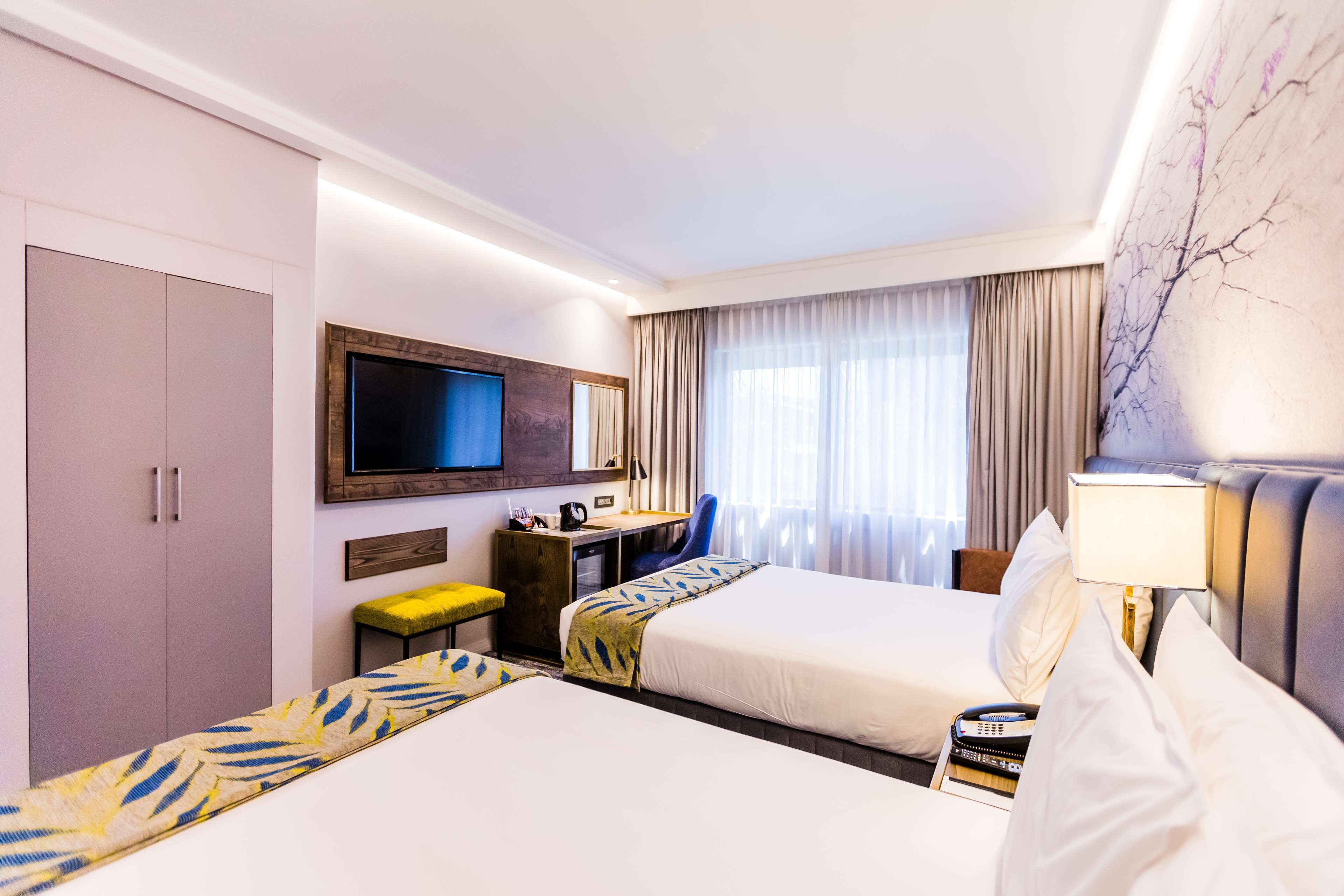 Habitación de hotel con dos camas, un escritorio con silla, televisión y un gran ventanal con cortinas.