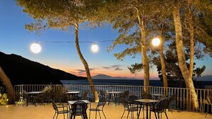 Une terrasse extérieure avec des tables et des chaises, une balustrade et une vue sur la mer et les montagnes.