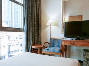 A hotel room with a bed, a desk with a television, a chair, a lamp, and a view of buildings through the window. - Classique Hotel (Singapore)