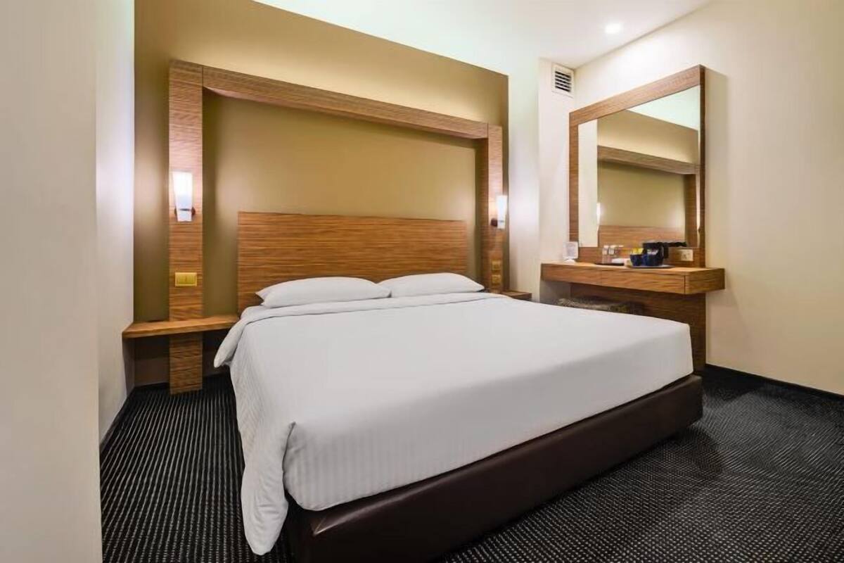 A hotel room with a bed, a wooden headboard, a nightstand, a mirror, and a small wooden dresser.