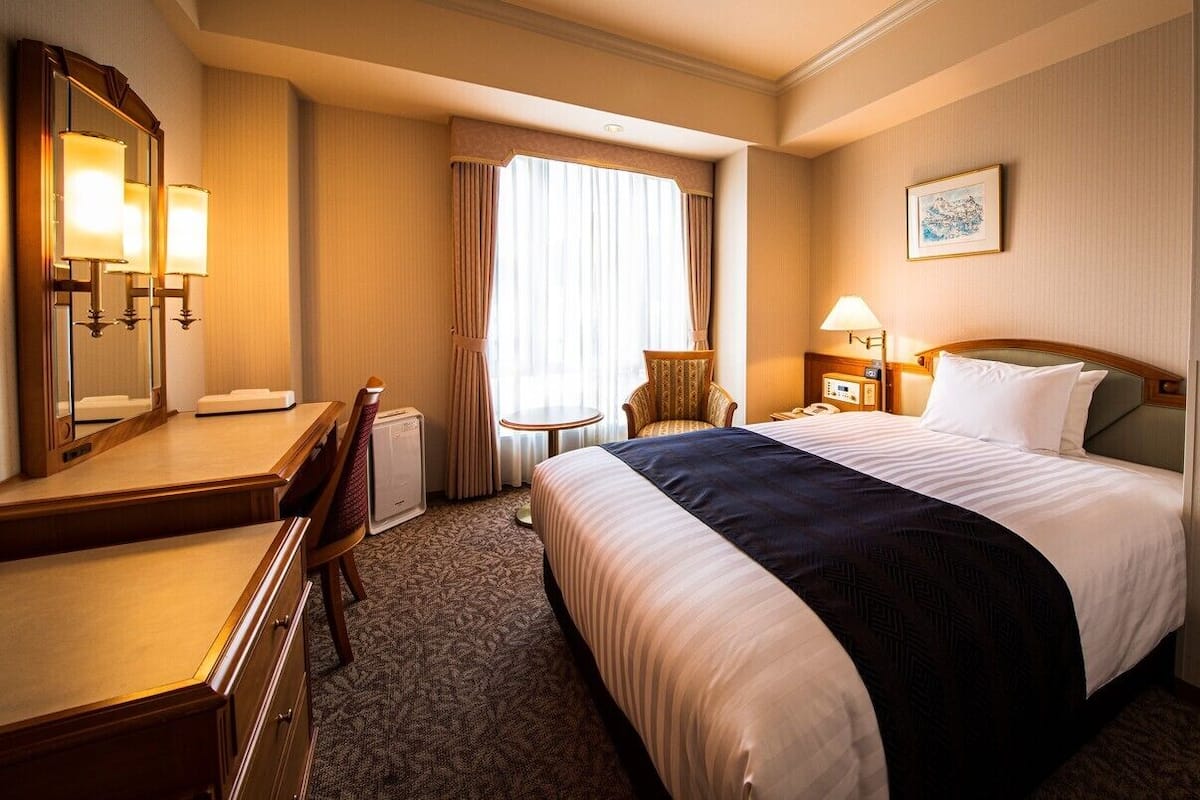 A hotel room with a bed, a desk, a chair, a lamp, a clock, and a painting on the wall.