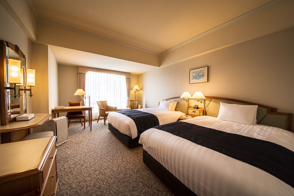 A hotel room with two beds, a desk, a chair, and a small table.
