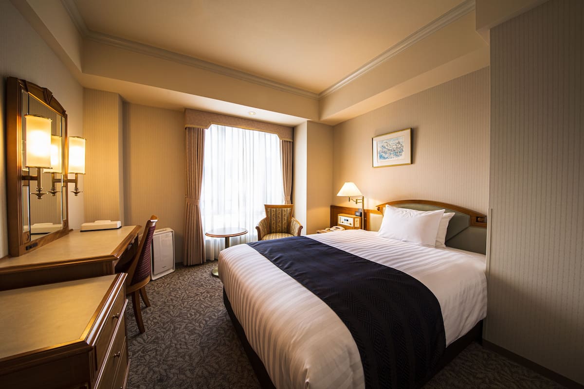 A hotel room with a large bed, a desk with a mirror, a chair, a small table, and a lamp.