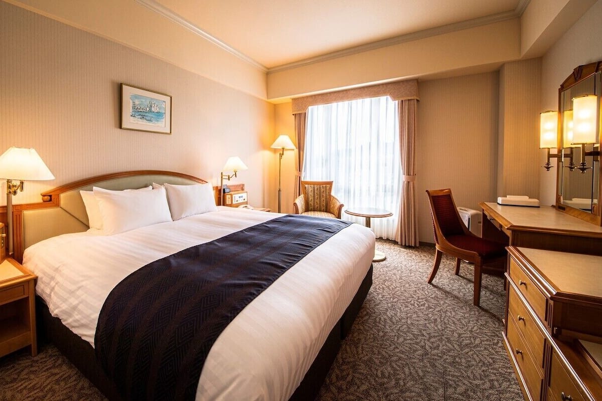 A hotel room with a large bed, bedside tables, a desk, a chair, a mirror, and a window with curtains.