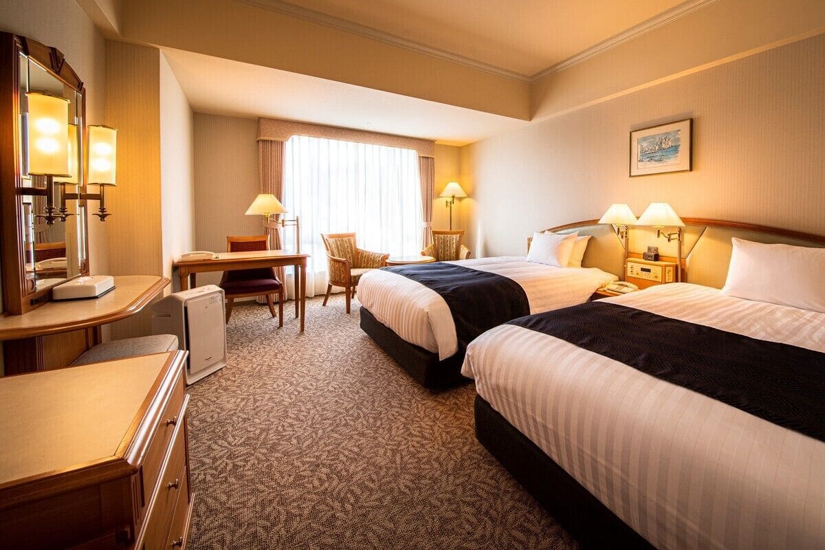 A hotel room with two beds, a desk, a chair, a lamp, and a painting on the wall.
