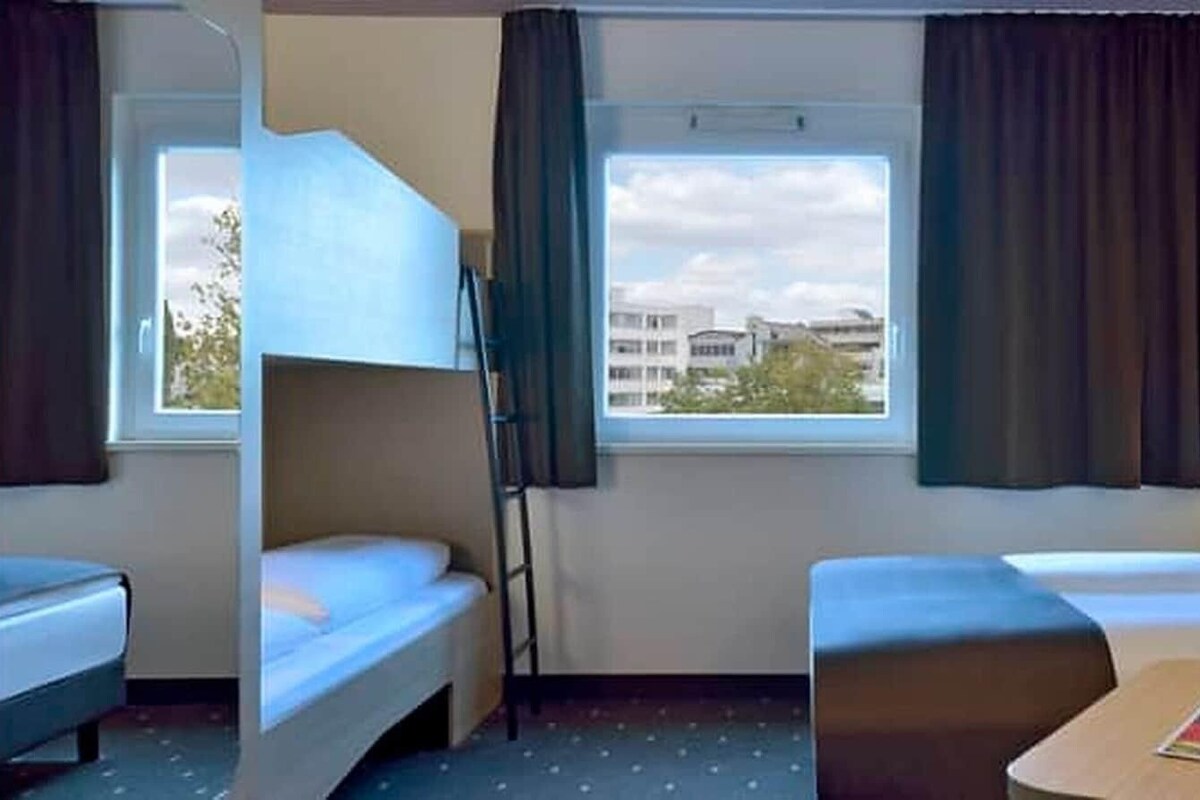 Habitación de hotel con dos camas, una ventana con cortinas, una mesita con una revista y un control remoto, y una lámpara fijada a la pared.