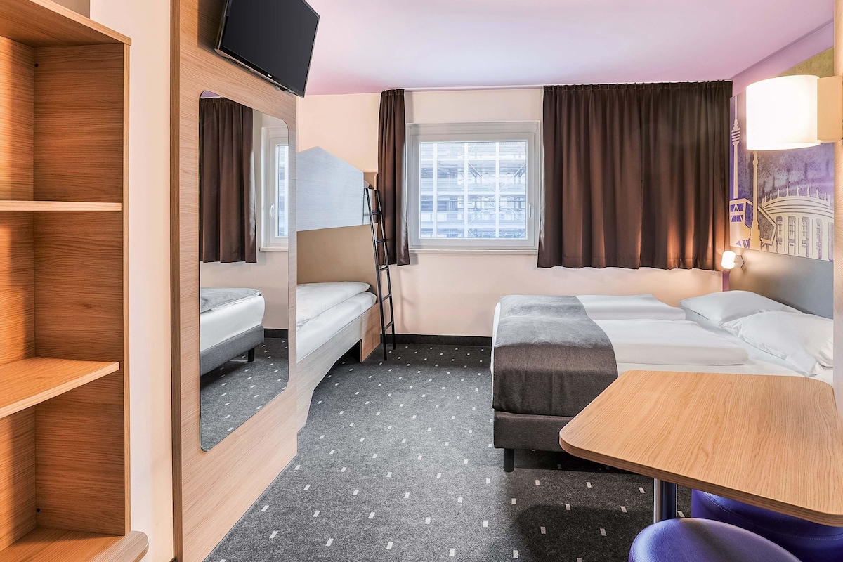 Une chambre d’hôtel avec un lit superposé, un bureau, une chaise, une télévision et une fenêtre avec des rideaux.