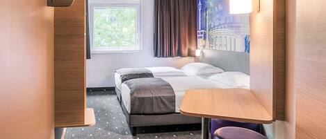 Habitación de hotel compacta con cama, una mesita, una silla, ventana con cortinas y una lámpara de pared.