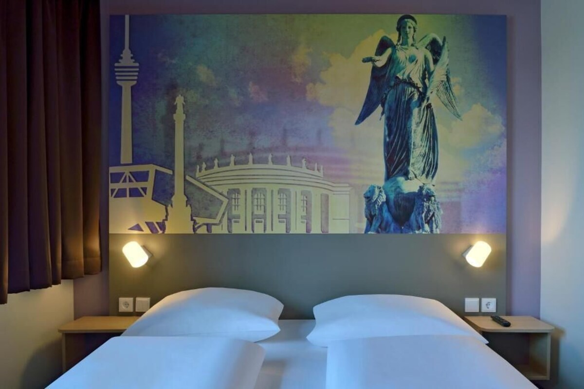 A hotel room with a bed, two bedside tables, and a large mural on the wall depicting a cityscape with a tower and an angel statue.