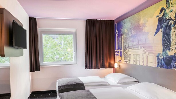 A hotel room with two beds, a TV, a window with curtains, and a mural on the wall. - B&B Hotel Stuttgart-Vaihingen (Stuttgart)