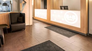 A modern reception area with a wooden reception desk, a seating area with a sofa and armchair, and a large mirror reflecting the interior. - B&B Hotel Stuttgart-Vaihingen (Stuttgart)