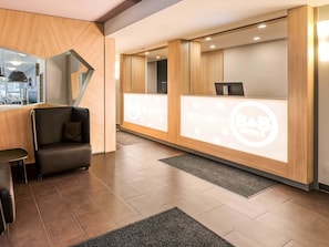 Un espace d’accueil moderne, doté d’un comptoir de réception en bois, d’un coin salon avec un canapé et un fauteuil, et d’un grand miroir qui reflète l’intérieur.