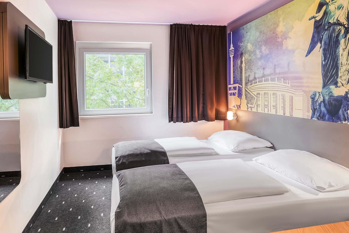 A hotel room with two beds, a TV, a window with curtains, and a mural on the wall.