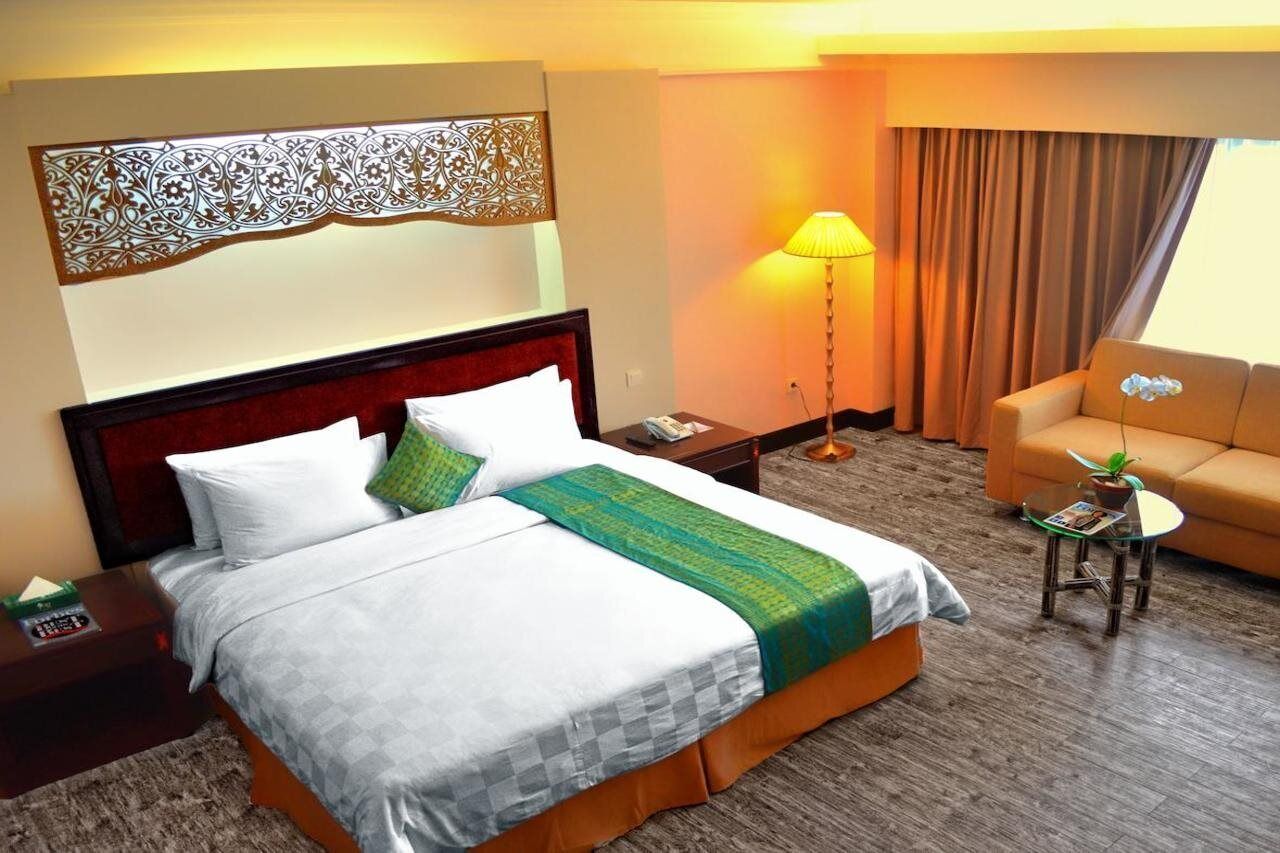 A hotel room with a bed, a sofa, a lamp, a small table with a plant, and a decorative wall piece.