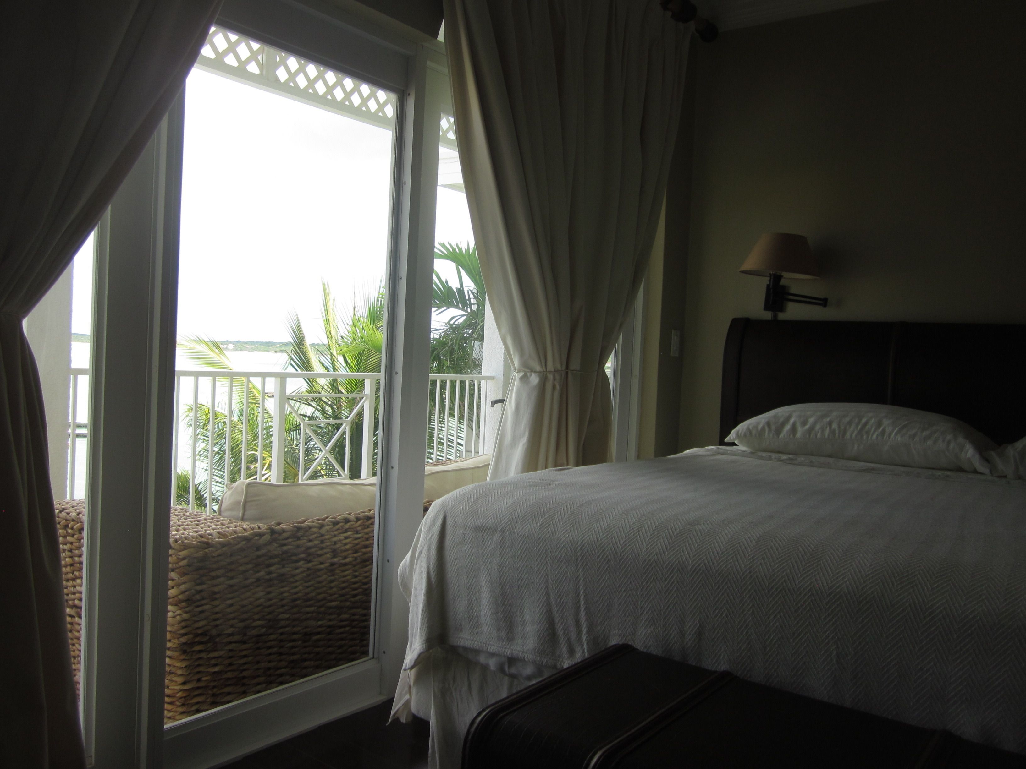 Un dormitorio con cama, ventana con cortinas, vista a un balcón con plantas y una lámpara en la pared.
