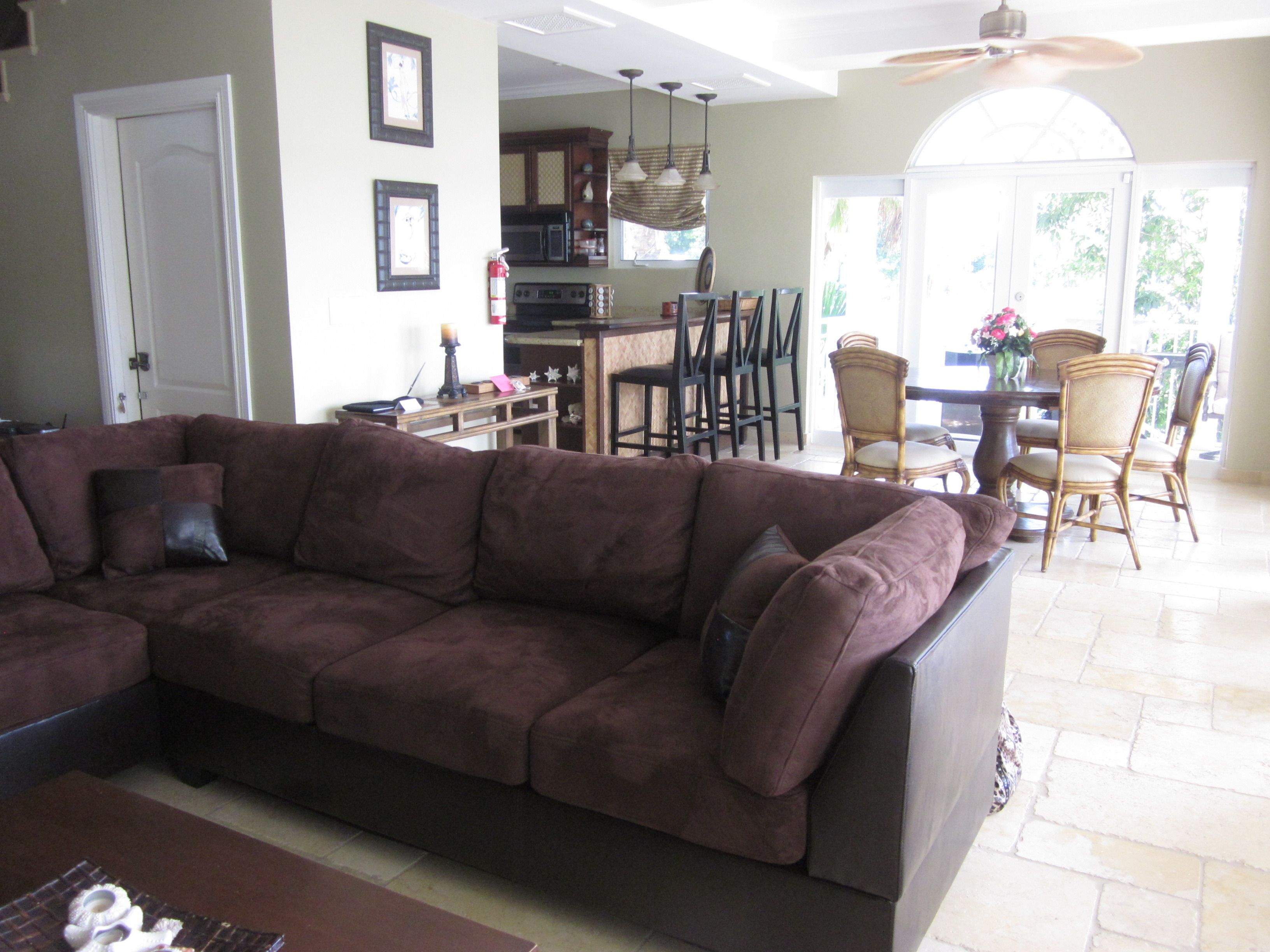 A living room with a large brown sectional sofa, a dining area with wooden chairs, and a kitchen in the background.