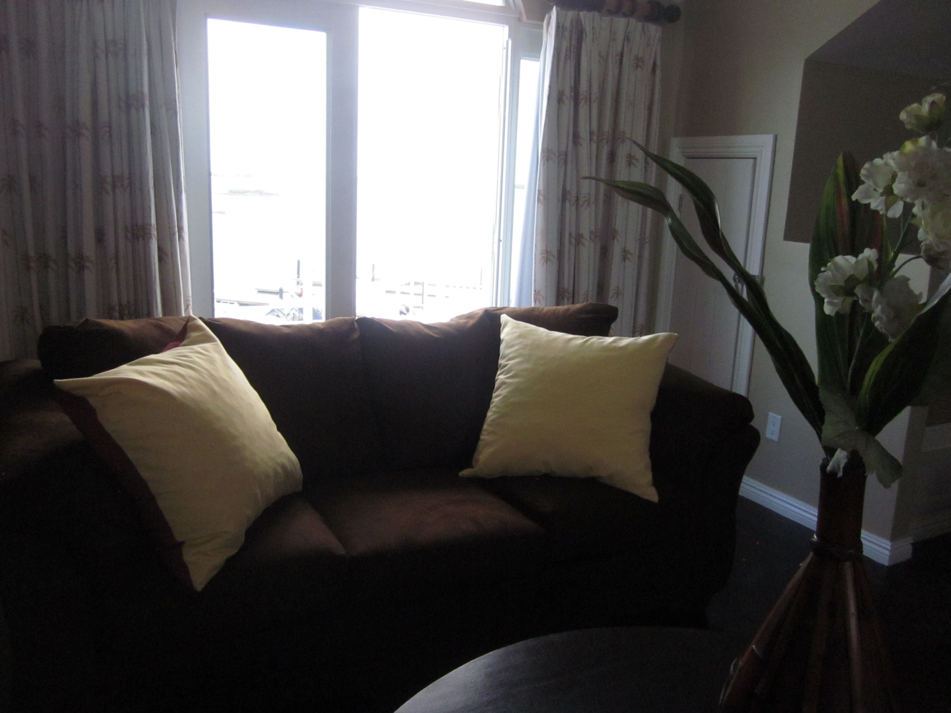A living room with a brown sofa, two yellow pillows, and a vase with white flowers.
