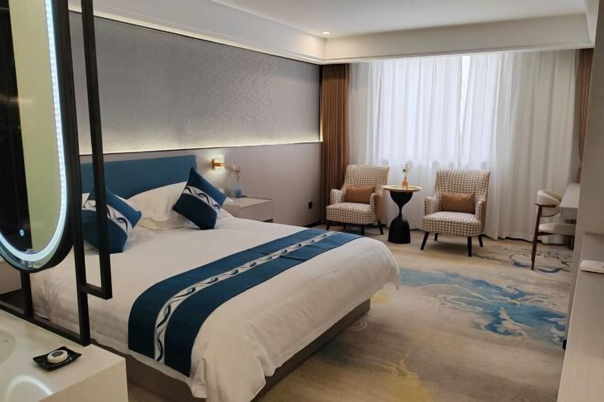 Ein Hotelzimmer mit einem großen Bett, zwei Sesseln, einem kleinen Tisch und einem großen Fenster mit Vorhängen.