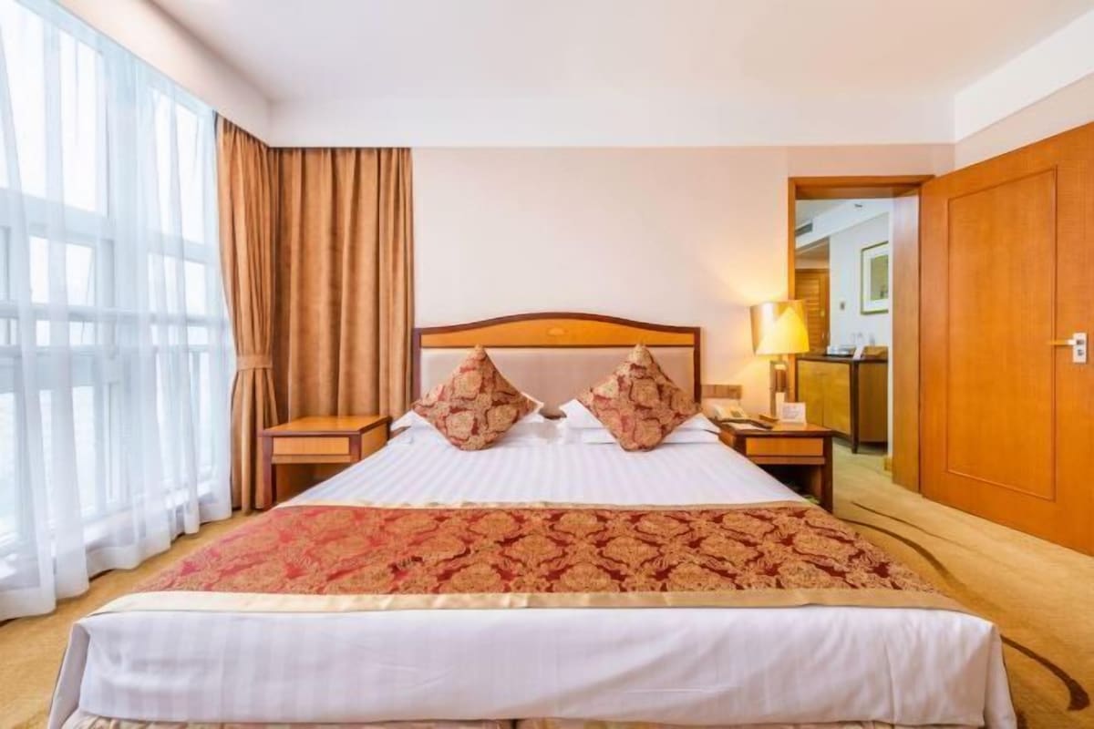 A hotel room with a large bed, two nightstands, a wooden headboard, and a view through a window with curtains.