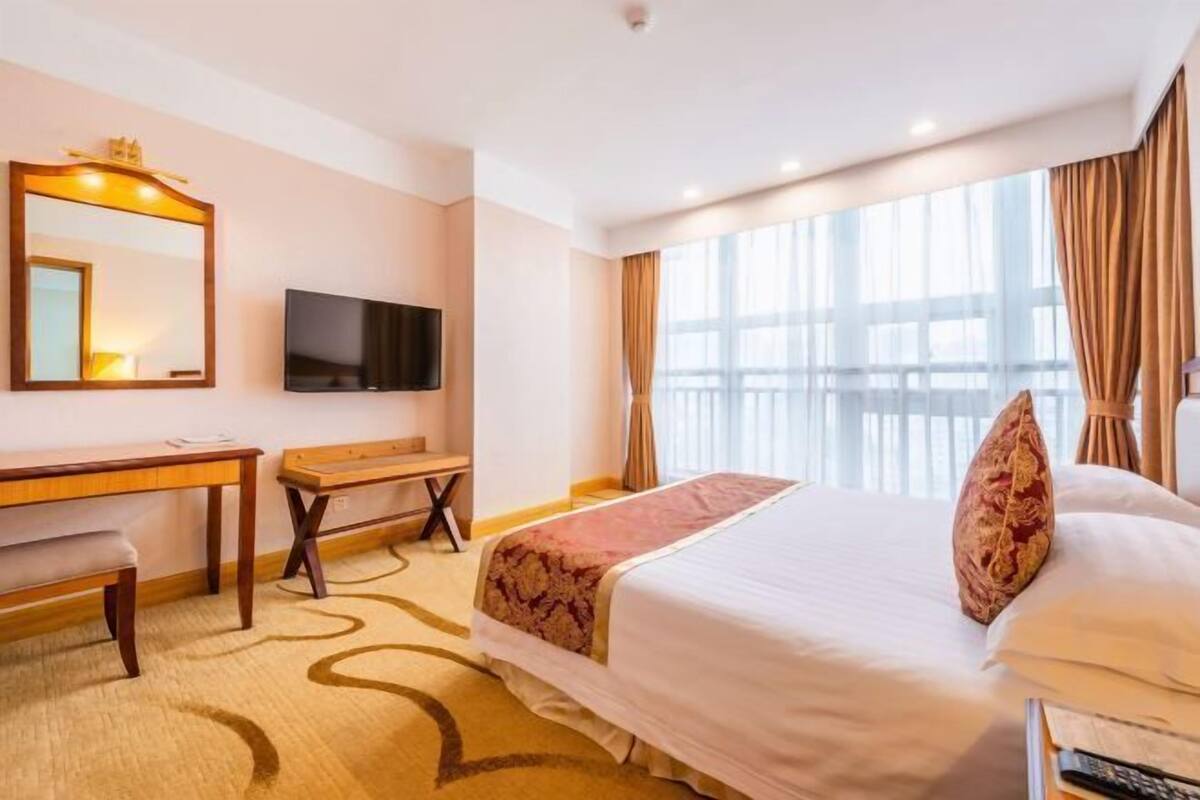 Ein Hotelzimmer mit Bett, Schreibtisch, Stuhl, Fernseher und einem großen Fenster mit Vorhängen.