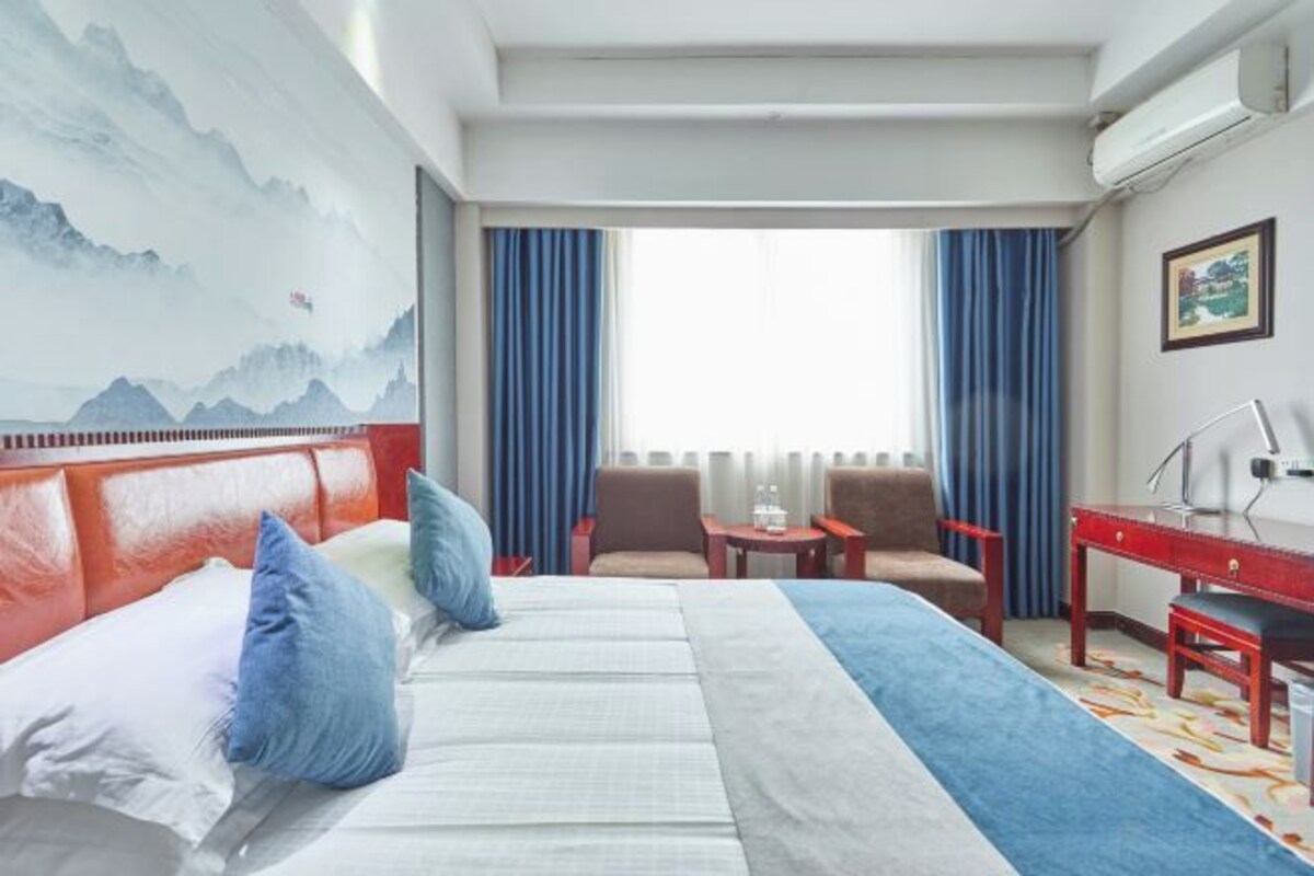 A hotel room with a bed, a desk, a chair, a small table, and a painting on the wall.